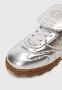 Hõbedane metallik tenniseking valgete paeltega ja kummist tallaga, millel on bränditud keelega flapp, millel on reljeefsed logo detailid.