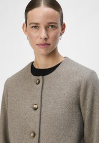 Beżowy płaszcz z wełny z okrągłym kołnierzem, czterema okrągłymi brązowymi guzikami i teksturowanym wykończeniem. Model nosi czarną koszulę wewnętrzną.