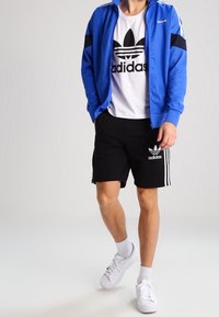 Blå zip-up idrottsjacka med svarta detaljer, vit Adidas t-shirt, svarta shorts med vita ränder och vita sneakers.
