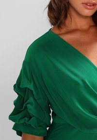 Grön klänning med djup V-ringning och böljande, volangärmar. Tyget verkar slätt och lätt, vilket förstärker dess flödande design.