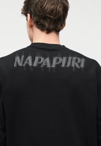 Άτομο με κοντά καστανά μαλλιά που φοράει μαύρο φούτερ με παραμορφωμένο γκρι κείμενο στο πάνω μέρος της πλάτης, σε απλό φόντο.