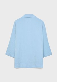 Veste bleu clair texturée à manches longues avec col, vue de dos sur un fond gris clair uni.