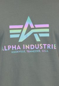 Pilkas medvilninis marškinėliai su holografiniu logotipu, kuriame yra „A“ ir trys horizontali juostos. Tekstas skelbia „ALPHA INDUSTRIE“ ir vietos detales.