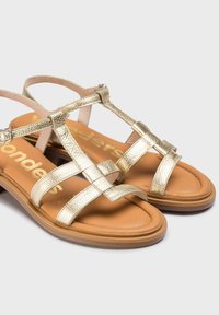 Sandales métalliques dorées avec des brides entrecroisées, une semelle intérieure rembourrée beige et une semelle basse texturée. Comprend une bride de cheville réglable.