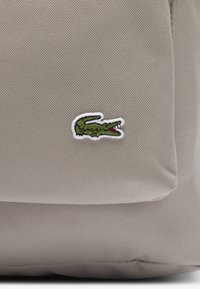 Beżowa tkanina o teksturowej powierzchni z małym haftowanym zielonym logo krokodyla z otwartą czerwoną paszczą.