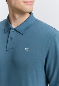 Polo skjorte i teal farvet stof med en struktureret overflade. Har en krave og to knapper, og viser et lille hvidt logo på brystet.