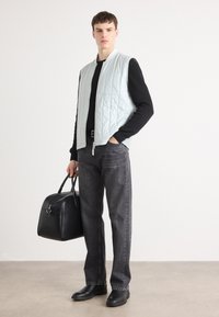 Colete acolchoado azul claro sem mangas sobre uma camisola preta de mangas longas, combinado com jeans cinzentos e botas de couro pretas, segurando uma bolsa preta.