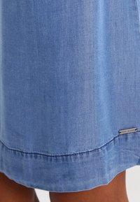 Denimrock in einem hellblauen Farbton, nahtloser Saum und metallisches Markenzeichen am unteren Ende. Weiche Textur mit einem leicht verblassten Finish.