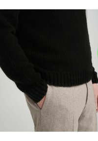 Schwarzer Strickpullover mit strukturierter Oberfläche, ausgestattet mit gerippten Bündchen und Saum. Kombiniert mit hellgrauen Hosen, die eine Taschenkante zeigen.