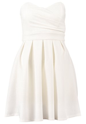 Frau in einem weißen, trägerlosen, plissierten Kleid, hält eine kleine rechteckige Clutch und trägt schwarze Sandalen mit hohen Absätzen, geht vorwärts.