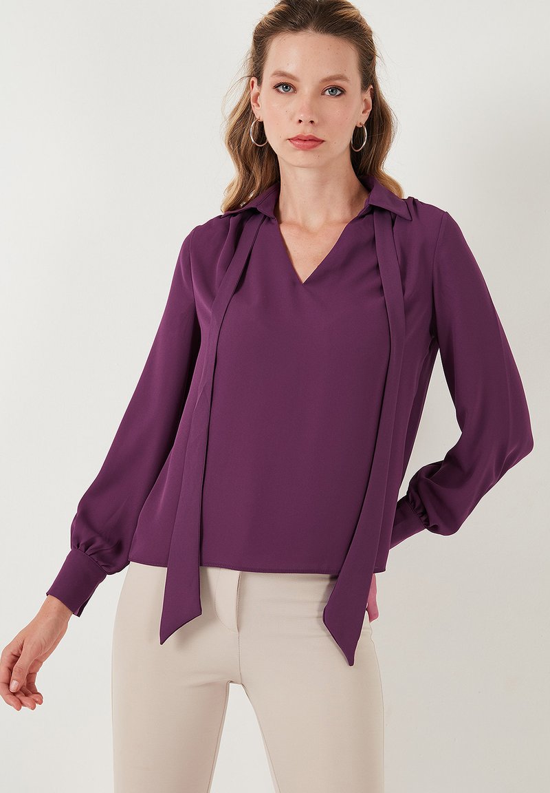 Blusa lila de manga larga con escote en V y collar estructurado, que presenta lazos fluidos en el frente y mangas fruncidas, hecha de una tela suave.