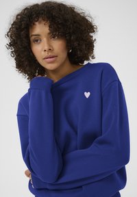 Modrá mikina s volným střihem, s ribovaným výstřihem a manžetami a malým růžovým srdíčkem vyšitém na hrudi. Hladká textura.