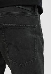Jeans de denim negro con un bolsillo trasero que presenta detalles de costura sutiles y una textura suave. Etiqueta parcialmente visible sobre la cintura.