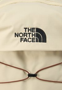 Μπεζ υφασμάτινο σακίδιο με υφή πλέγματος, με έντονο μαύρο κεντημένο λογότυπο και καφέ ελαστικό κορδόνι για ασφαλές κλείσιμο.