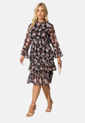 Kleid mit Blumenmuster auf schwarzem Hintergrund, mit Rüschenlagen, langen transparenten Ärmeln und hohem Ausschnitt. Das Model hält eine hellfarbige Clutch.