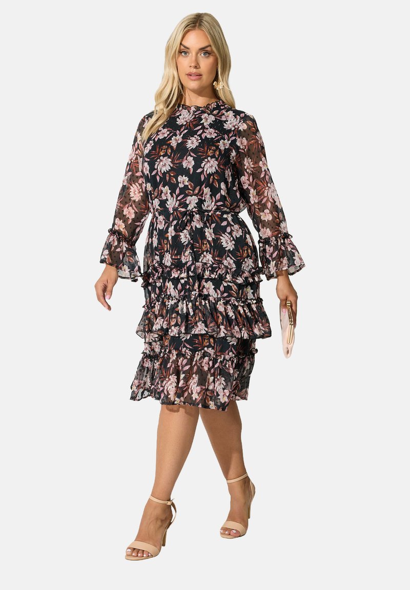 Klänning med blommigt mönster på svart bakgrund, volanger, långa genomskinliga ärmar och hög krage. Modellen håller en ljusfärgad clutchtvå.