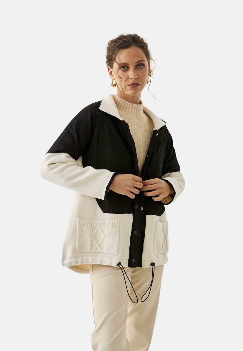 Mujer vestida con una chaqueta de lana negra y crema con bolsillos frontales, ajustando los botones, combinada con pantalones crema y pendientes de aro dorados.