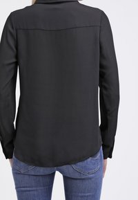 Blusa di seta nera con una texture liscia, maniche lunghe e orlo curvo, dettagliata con una cucitura sulla spalla posteriore per offrire struttura.