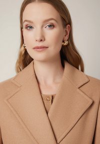 Beżowy płaszcz wełniany z wyciętym kołnierzem, z ozdobnym złotym guzikiem przy dekolcie. Modelka nosi duże złote kolczyki w kształcie kółek.