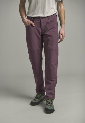 Person trägt pflaumenfarbene Hose mit mehreren Taschen und graue Wanderschuhe, steht vor neutralem einfarbigem Hintergrund.