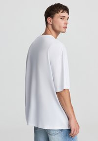 T-shirt bianca oversize con scollatura rotonda e maniche corte, realizzata in tessuto morbido, indossata con jeans azzurri chiari, vista da dietro.