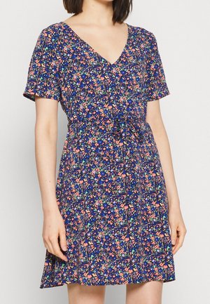Woman wearing a short-sleeve, V-neck dress with a blue background and small pink, orange, and green floral pattern, tied at the waist.