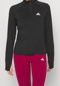 Schwarzes, langärmeliges Sportoberteil mit einem halben Reißverschluss und einem kleinen weißen Adidas-Logo; kombiniert mit engen, knallrosa Leggings mit weißen Streifen.