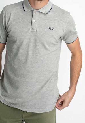 Homme portant un polo à manches courtes gris clair avec deux boutons et des bordures foncées au col et aux manches, associé à un pantalon vert.