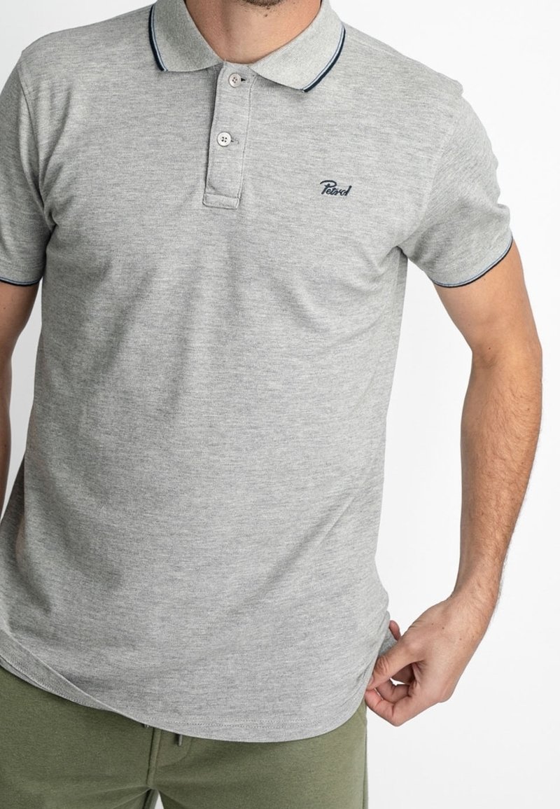 Homme portant un polo à manches courtes gris clair avec deux boutons et des bordures foncées au col et aux manches, associé à un pantalon vert.