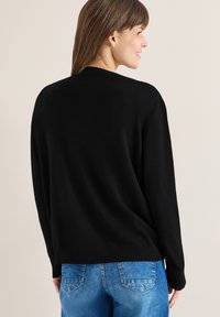 Femme de profil portant un pull noir à manches longues et un jean denim bleu, souriant devant un fond clair uni.