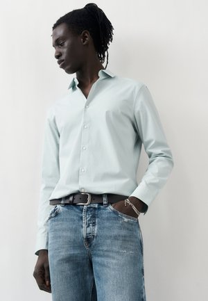 Mężczyzna o ciemnej karnacji i dredach, ubrany w jasnomiętową koszulę zapinaną na guziki, wpuszczoną w wyblakłe niebieskie dżinsy z czarnym paskiem i srebrną bransoletką.