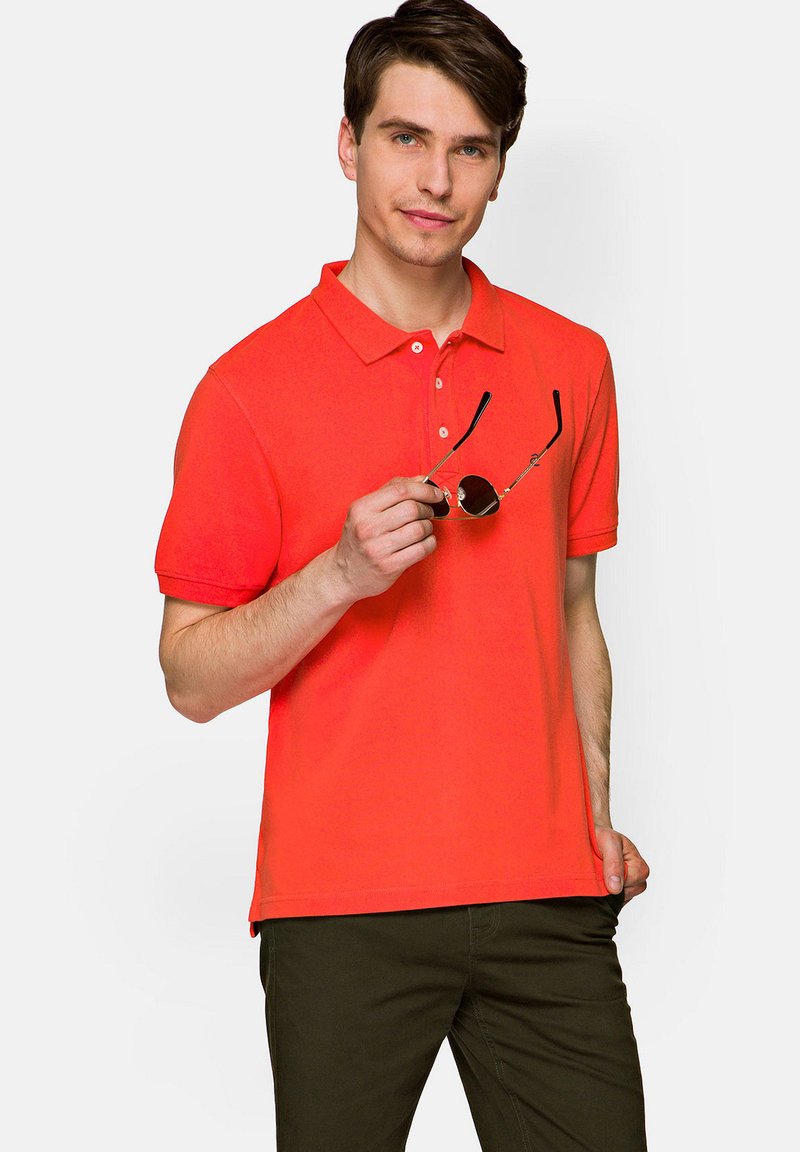 Koszulka polo z krótkim rękawem w jaskrawym pomarańczowym kolorze z kołnierzykiem i trzema guzikami, w zestawieniu z ciemnozielonymi spodniami; trzymająca okrągłe okulary przeciwsłoneczne.