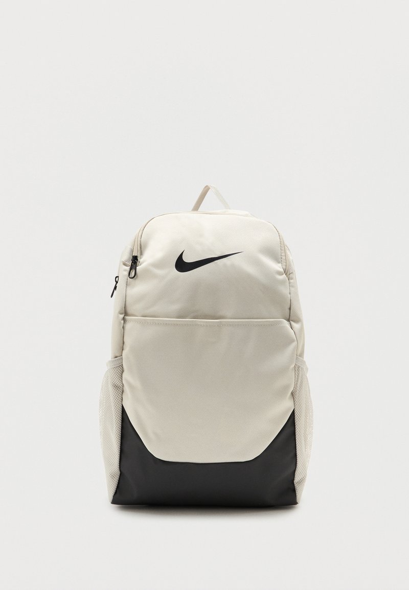 Sac à dos Nike beige clair et noir avec poche avant à fermeture éclair, poches latérales en mesh et poignée supérieure sur un fond uni.