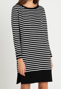Vestido de mangas largas a rayas en blanco y negro con un escote redondeado, que presenta un dobladillo negro sólido y aberturas laterales para mayor comodidad al moverse.