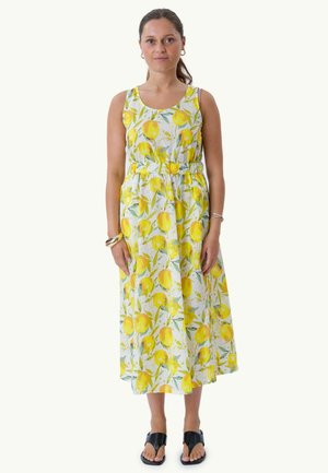 Femme debout portant une robe midi blanche sans manches avec un motif citron jaune et feuille verte et des sandales noires à bout ouvert.