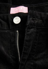 Κοντινή λήψη μαύρου κοτλέ παντελονιού με ασημί φερμουάρ και κουμπί, με ροζ ετικέτα που γράφει JJXX Girl στη μέση.