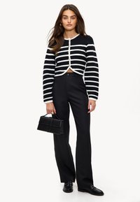 Cardigan court à rayures noires et blanches avec un col rond, fermeture par boutons et manches longues, associé à un pantalon noir évasé et une pochette.