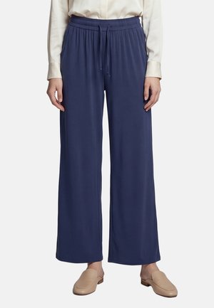 Pantalon large bleu marine avec taille élastique et cordon de serrage, porté avec un chemisier crème et des chaussures beiges à enfiler, montré de la taille aux pieds.