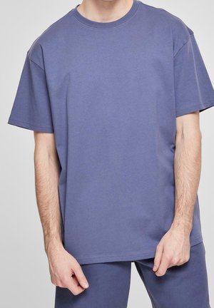 Mann trägt ein schlichtes blaues T-Shirt mit kurzen Ärmeln und dazu passende Hosen, steht vor einem einfarbigen hellen Hintergrund.