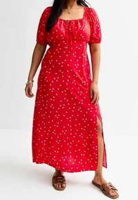 Rotes Maxikleid mit gepunktetem Muster, Puffärmeln, quadratischem Ausschnitt und seitlichem Schlitz. Hergestellt aus leichtem Stoff. Begleitet von braunen flachen Sandalen.