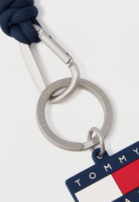 Ναυτικό μπλεγμένο ύφασμα μπρελόκ που διαθέτει μεταλλικό ασημένιο καραμπίνερ και μια ορθογώνια ετικέτα με σχέδιο σε κόκκινο, λευκό και μπλε χρώμα, με ανάγλυφο κείμενο.