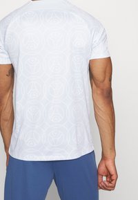 Camiseta atlética blanca con un sutil patrón circular, mangas cortas y cuello redondeado, combinada con pantalones cortos azules.