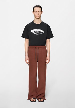 Férfi fekete pólóban, amelyen fehér szem grafika található, barna, széles szárú, zsinóros nadrágban és fekete papucsban, egyszínű háttér előtt áll.