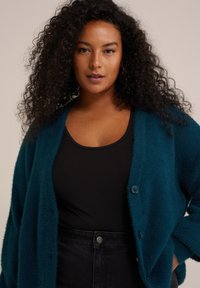 Teal, fluffig cardigan med lös passform, som har två knappar och ribbad textur, bärs över en svart linne och en högmidjad svart kjol.
