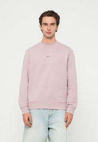 Muž oblečený v svetloružovej mikine BOSS a svetlomodrých džínsoch stojaci na bielom pozadí.