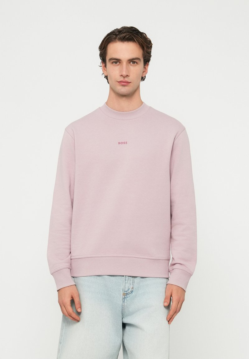 Muž oblečený v svetloružovej mikine BOSS a svetlomodrých džínsoch stojaci na bielom pozadí.