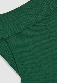 Grünes Strickkleidungsstück mit einem gerippten Taillenbund und einem strukturierten Körper mit vertikalen Linien. Der Stoff wirkt weich und dehnbar.