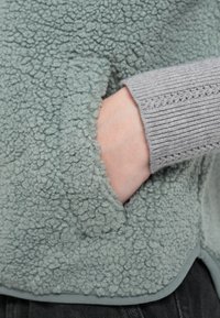 Pulôver de lã polar macia e texturada em verde, com uma mão num bolso; manga cinzenta canelada com um padrão delicado perto do punho.