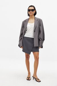 Blazer oversized gris avec des manches larges, top blanc en débardeur et jupe enveloppante gris foncé. Les accessoires comprennent des lunettes de soleil noires et des talons fins noirs.