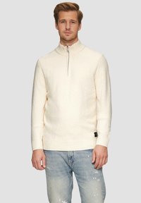 Sweter w kolorze kremowym z połową rozpięcia, wykonany z fakturowanej tkaniny, z ściągaczem przy kołnierzu i dolnej krawędzi, z małą czarną etykietą po lewej stronie na dole.